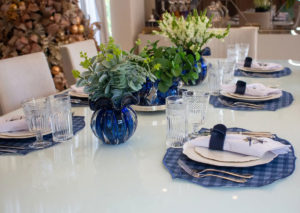 Mesa posta com decoração elegante em tons de azul e branco, incluindo vasos de vidro azul com arranjos verdes, pratos com jogos americanos xadrez e copos de vidro trabalhado.