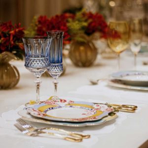 Mesa posta com pratos estampados, copos de cristal azul lapidados e talheres dourados, decorada com arranjos de flores vermelhas em vasos de vidro âmbar.