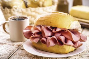 Sanduíche de pão francês recheado com fatias de mortadela, servido em um prato branco, acompanhado de uma xícara de café quente sobre uma mesa rústica, com cesta de pães e queijo ao fundo.