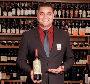 Homem sorridente, vestido com terno preto e camisa vermelha, segurando uma garrafa de vinho e apresentando-a em uma adega ou loja de vinhos, com prateleiras repletas de garrafas ao fundo.