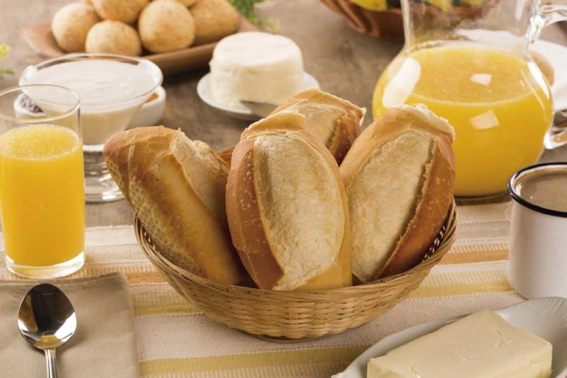 Mesa de café da manhã com cesta de pães franceses, suco de laranja, café, manteiga, queijo branco e pães de queijo, sobre uma toalha bege listrada, criando um ambiente acolhedor.