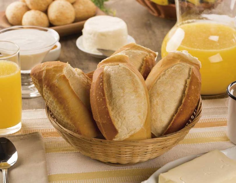 Mesa de café da manhã com cesta de pães franceses, suco de laranja, café, manteiga, queijo branco e pães de queijo, sobre uma toalha bege listrada, criando um ambiente acolhedor.